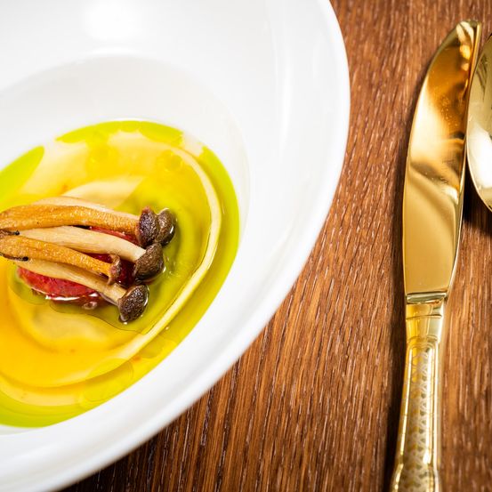 Eine weiße Schüssel mit gelber Flüssigkeit und einigen Pilzen, neben einem goldenen Messer auf einem Holztisch.