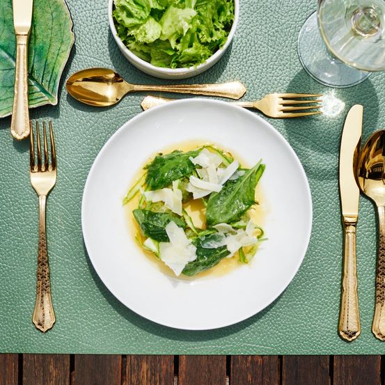 Ein weißer Teller mit einem grünen Spinatgericht auf einem grünen Platzset, mit Besteck und einer Schüssel Salat.