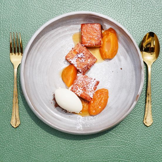 Ein Gericht aus French-Toast-Stücken, die mit Puderzucker bestreut sind, auf einem Teller mit Aprikosen und einer Kelle Sahne. Es wird mit goldfarbenen Besteck auf einer grünen Oberfläche serviert.