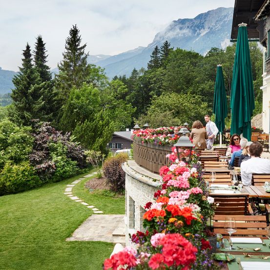 Eine malerische Außengastronomie mit Tischen und Stühlen, umgeben von Blumen, Grünpflanzen und einer wunderschönen Bergsicht in der Ferne.