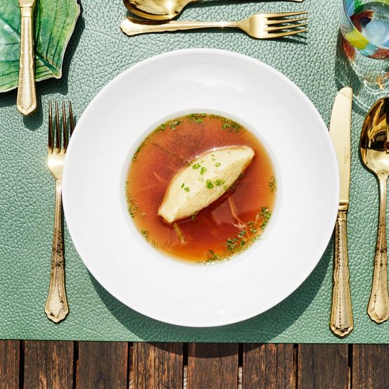 Ein weißer Teller enthält eine Suppe mit einem zentralen Fleischstück, garniert mit Kräutern, auf einem grünen Platzset mit Besteck.