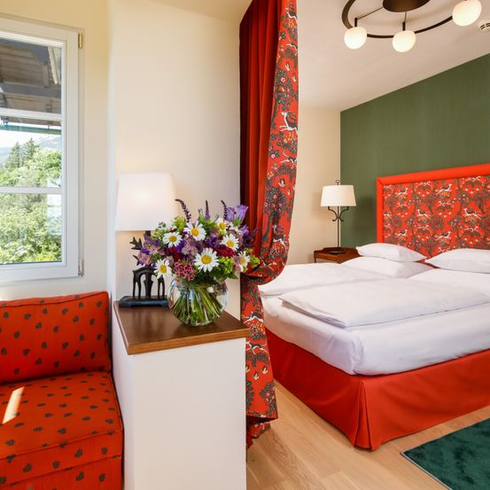 Ein gemütliches Schlafzimmer mit einem Bett mit weißen Laken und einem roten Kopfteil. Ein Vasen mit bunten Blumen steht auf einem Tisch neben einem roten Vorhang.