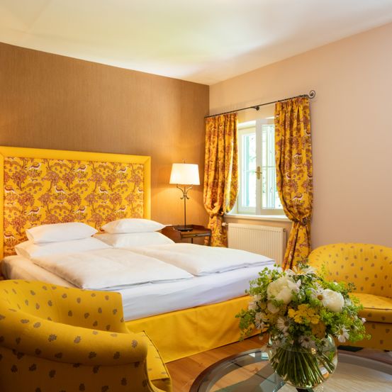 Ein gemütliches Schlafzimmer mit einem Doppelbett, geschmückt mit weißen Kissen und einem gelben Kopfteil. Gelbe Vorhänge hängen über einem Fenster, und eine Lampe steht auf einem Nachttisch.