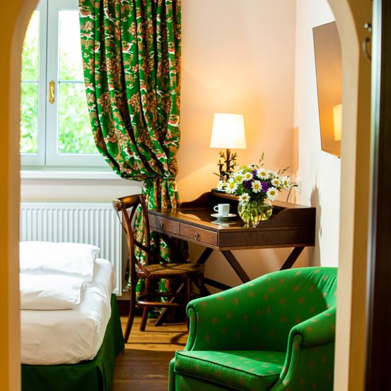 Ein Schlafzimmer mit einem Bett, einem Holzschreibtisch, einer Lampe und einem Stuhl. Ein grüner Stuhl steht neben dem Schreibtisch. Das Zimmer hat ein Fenster mit Vorhängen. Der Boden ist aus Holz.