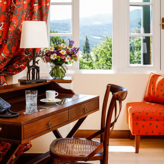 Ein Zimmer mit einem Holzschreibtisch, einem braunen Stuhl und einem Telefon. Ein Lampenschirm mit einem Blumenvase steht auf dem Schreibtisch. Das Zimmer hat ein Glasfenster mit Blick auf Berge.