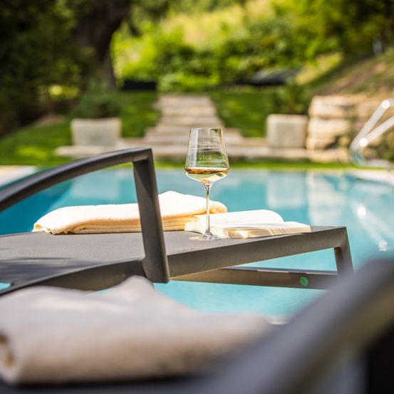 Ein Glas Wein steht auf einer Liege am Pool, mit Handtüchern und einem Buch in der Nähe, umgeben von einer ruhigen Außenumgebung mit Grünflächen und Treppen.