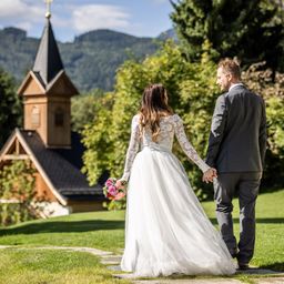Eine Braut in einem weißen Kleid und ein Bräutigam in einem grauen Anzug halten Händchen und gehen auf eine kleine Kirche mit einem Turm zu, die von Grün umgeben ist.
