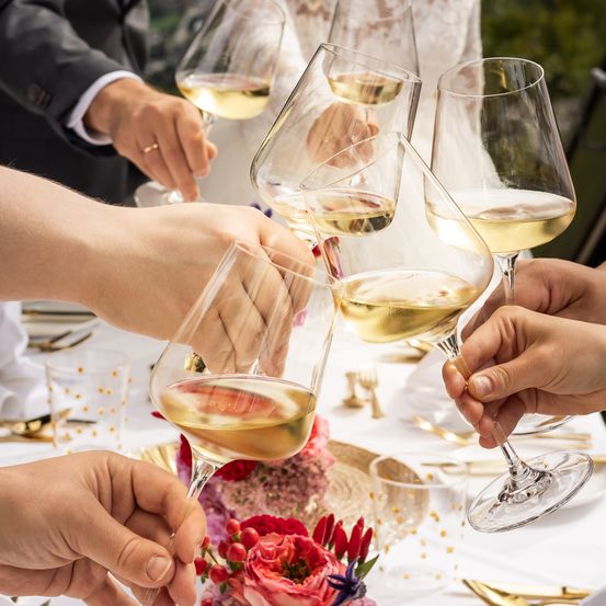 Eine Gruppe von Menschen stößt mit Gläsern Weißwein an einem Tisch mit Blumen und Besteck an.