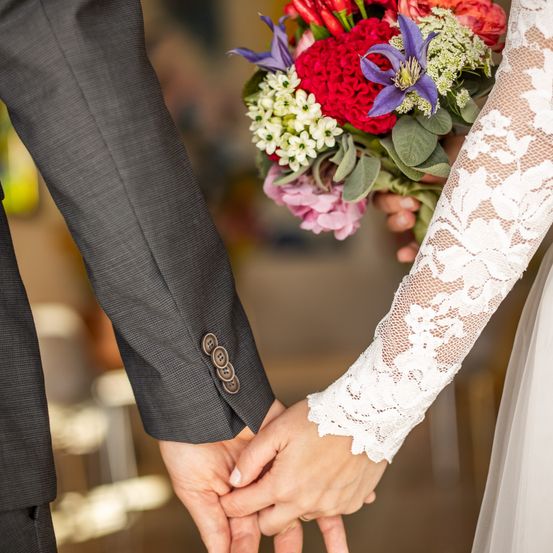 Ein Brautpaar hält bei einer Hochzeitszeremonie Händchen und hat einen farbenfrohen Blumenstrauß dazwischen.