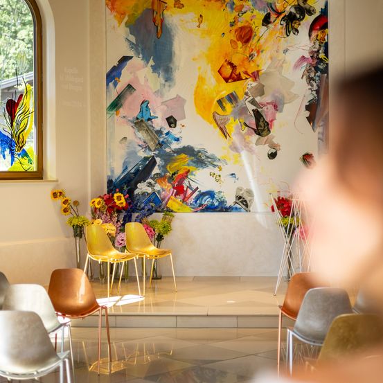 Ein farbenfrohes abstraktes Gemälde ist an einer weißen Wand aufgehängt, mit einem Fenster und Blumen in Vasen in der Nähe. Stühle sind in Reihen angeordnet, und eine Person ist im Vordergrund leicht verschwommen.