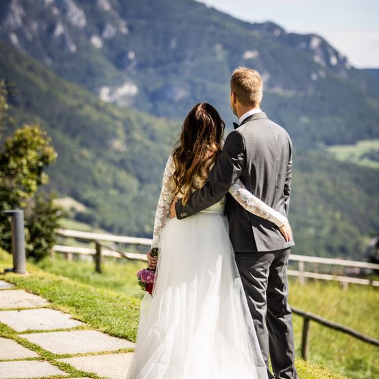 Ein frisch verheiratetes Paar, die Braut in einem weißen Kleid und der Bräutigam in einem grauen Anzug, steht auf einem Rasenhügel mit einem Holzzaun und blickt auf eine Berglandschaft.