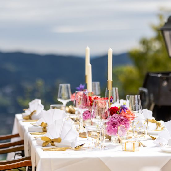 Eine wunderschön gedeckte Tischlandschaft im Freien mit weißer Tischdecke, Weingläsern, Blumen und Kerzen. Berge und ein klarer Himmel im Hintergrund.