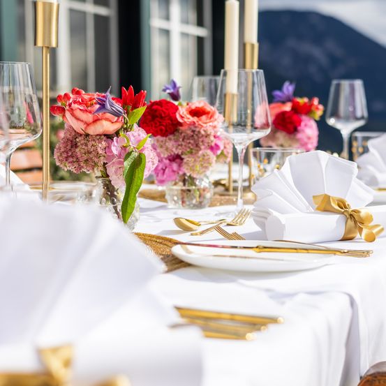 Ein Tisch ist mit Blumen, Weingläsern, Besteck und Servietten gedeckt. Es gibt Kerzen und eine Aussicht auf Berge im Hintergrund.