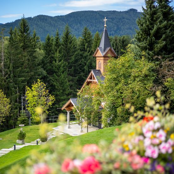 Eine kleine Holzkapelle mit einem Turm und einem Kreuz steht auf einem üppig grünen Hügel, umgeben von Bäumen und Blumen. In der Ferne sind Berge sichtbar.