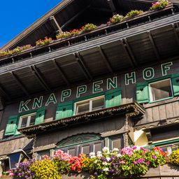 Eine Nahaufnahme eines Holzgebäudes mit dem Wort Knappenhof in grünen Buchstaben. Das Gebäude hat grüne Fensterläden und einen Balkon mit bunten Blumen.