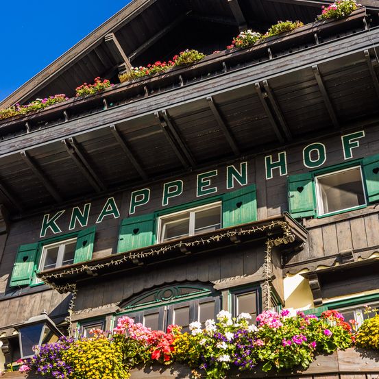 Eine Nahaufnahme eines Holzgebäudes mit dem Wort Knappenhof in grünen Buchstaben. Das Gebäude hat grüne Fensterläden und einen Balkon mit bunten Blumen.