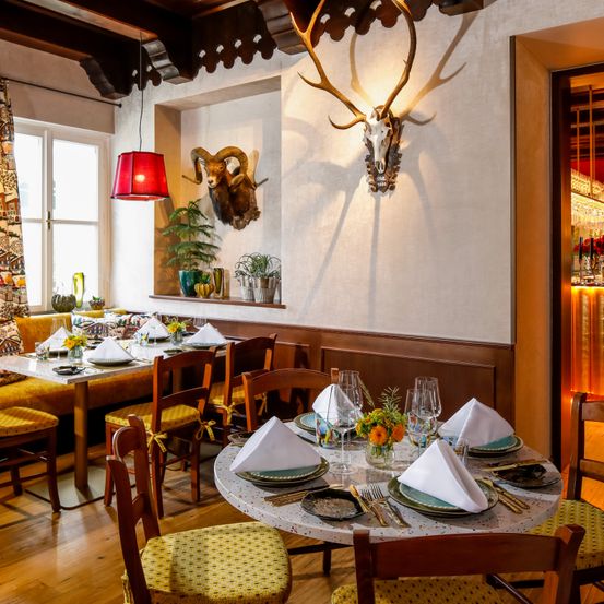 Ein Essbereich mit Tisch und Stühlen, Holzboden und zwei montierten Widderköpfen. Es gibt Topfpflanzen auf einem Regal, eine rote Lampe und ein Fenster.