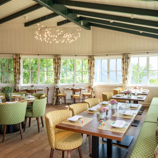 Ein Innenrestaurant mit Holzböden und Balken, ausgestattet mit Kronleuchtern, großen Fenstern und Tischen mit Blumenarrangements.