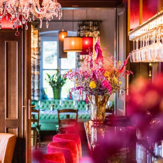 Ein elegantes Zimmer mit einer goldenen Vase mit bunten Blumen auf dem Tisch, einer Kristallleuchte und einem Fenster mit einer grünen Couch und Stühlen.