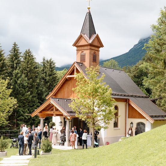 Eine Gruppe von Menschen steht vor einer kleinen Kapelle mit einem Turm, umgeben von Bäumen und Bergen.