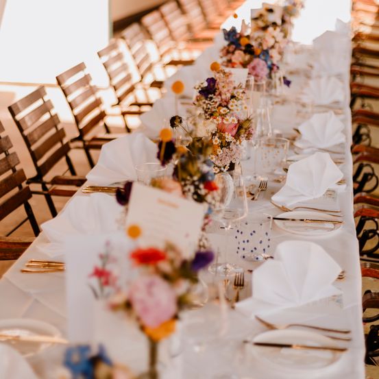 Ein langer Tisch für eine formelle Veranstaltung mit Blumensträußen, gefalteten Servietten, Gläsern und Besteck. Holzstühle sind um den Tisch herum angeordnet.