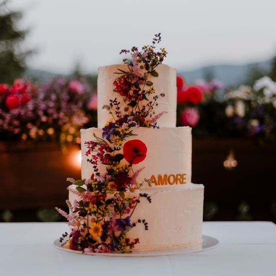 Ein dreistöckiger Hochzeitskuchen mit weißer Glasur, verziert mit verschiedenen Blumen und einer roten Mohnblume, steht auf einem weißen Tisch. Das Wort 'Amore' ist in Gold auf dem Kuchen geschrieben.