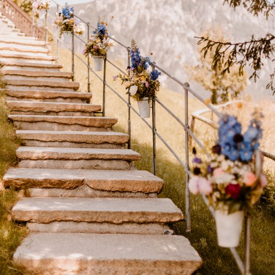 Steinstufen mit Metallgeländern sind mit weißen Blumentöpfen geschmückt und stehen vor einer Bergkulisse.