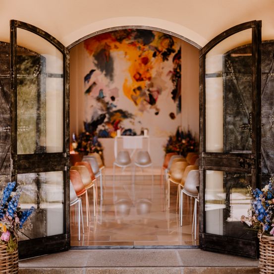 Eine offene Tür mit Metallscharnieren, die in einen Raum mit einer abstrakten Malerei an der Wand führt, und Stühle in einer Reihe aufgestellt. Es gibt Vasen mit Blumen auf den Seiten.