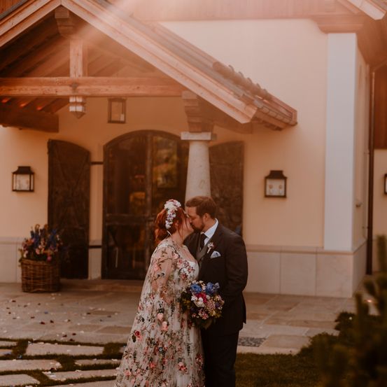 Ein Brautpaar küsst sich vor einem rustikalen Gebäude, umgeben von Blumen und Konfetti.