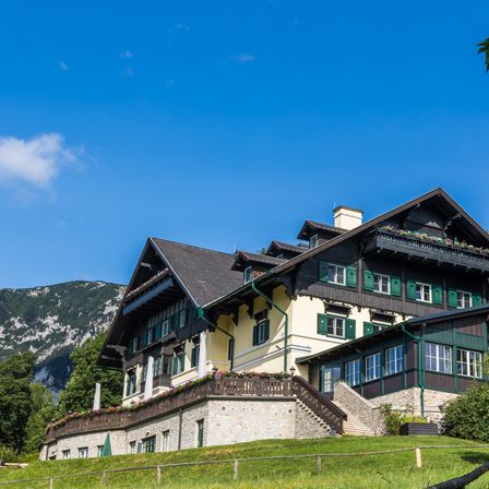 Ein großes Haus mit grünem Dach und weißen Wänden ist von einem grünen Rasen und Bäumen umgeben. Im Hintergrund befinden sich Berge.