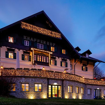 Das Knappenhof Hotel ist nachts beleuchtet. Es hat viele Fenster und einen Balkon mit Lichtern. Das Wort Knappenhof ist oben geschrieben.