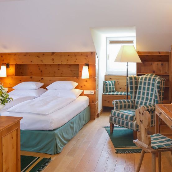 Ein Schlafzimmer mit Holzbett, grüner Bettdecke, Kissen und einem Holzschreibtisch mit Stuhl. Es gibt einen Stuhl mit Kissen und eine Lampe an der Seite.