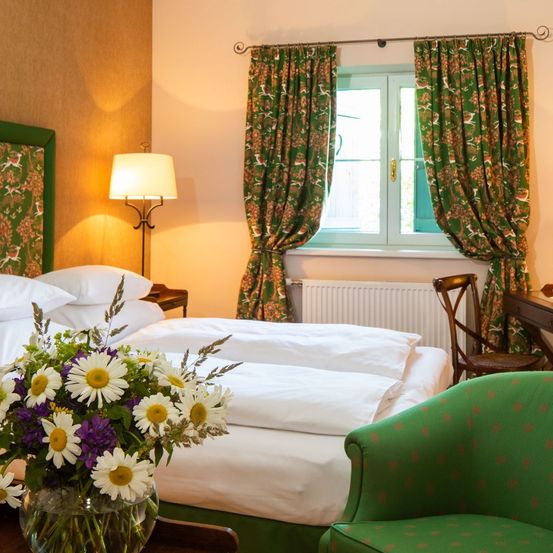 Ein Schlafzimmer mit einem Bett, einer Lampe und einem Fenster mit grünen Vorhängen. Auf dem Bett steht eine Blumenvase und ein Stuhl steht am Fenster.