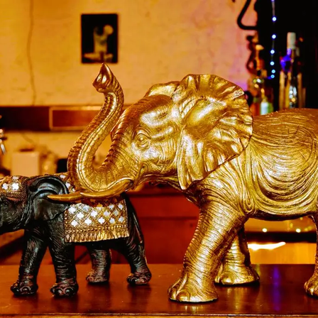 Zwei goldene Elefantenfiguren stehen auf einem Holztresen. Ein Elefant ist golden, während der andere schwarz mit goldenen Akzenten ist. Dahinter ist eine Wand mit einem Bilderrahmen.