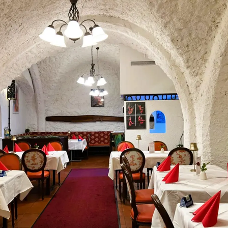Ein Restaurant mit gewölbten Decken hat Tische mit roten Servietten und beleuchteten Kronleuchtern. Die Wände sind aus Stein und es gibt einen roten Teppich. In der Ferne sind eine Bank und ein Bilderrahmen zu sehen.