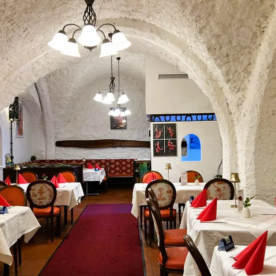 Ein Restaurant mit gewölbten Decken hat Tische mit roten Servietten und beleuchteten Kronleuchtern. Die Wände sind aus Stein und es gibt einen roten Teppich. In der Ferne sind eine Bank und ein Bilderrahmen zu sehen.