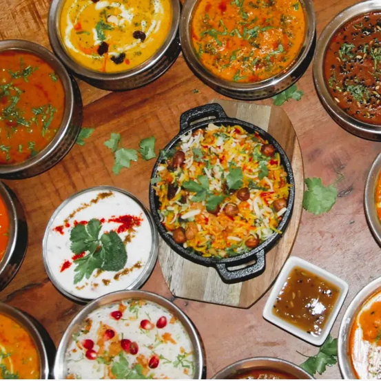 Auf einem Holztisch sind verschiedene indische Gerichte zu sehen, darunter Biryani, Curry und Chutney, garniert mit Kräutern und Gewürzen.