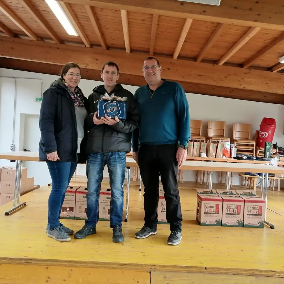 Drei Personen stehen auf einer Bühne in einem Saal. Ein Mann hält eine blaue Box. Ein anderer Mann lächelt. Eine Frau lächelt. Tische und Stühle stehen hinter ihnen. Kisten liegen auf dem Boden.