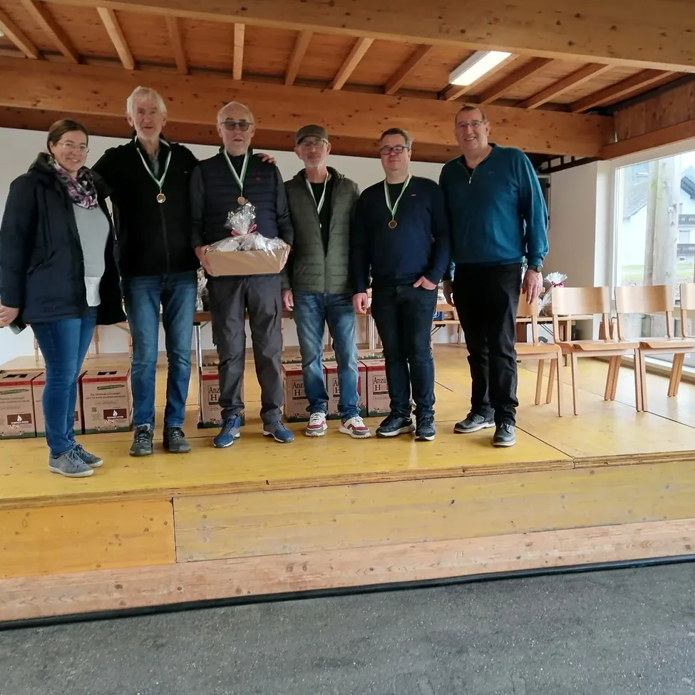 Sechs Personen stehen auf einer Bühne, wahrscheinlich bei einer Indoor-Veranstaltung, mit Brillen und Medaillen. Ein Mann hält eine Schachtel mit einem Geschenk darin. Dahinter stehen Holzstühle und Lagerboxen. Die Bühne hat einen Holzboden. Der Raum hat Holzbalken an der Decke.