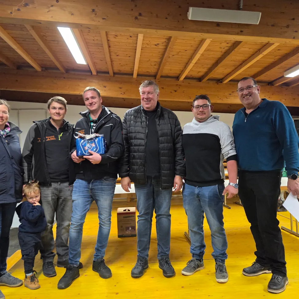 Eine Gruppe von Menschen, einschließlich eines Kindes, steht in einem Raum mit gelbem Boden und lächelt. Eine Person hält eine blaue Box. Sie sind in Winterkleidung gekleidet.