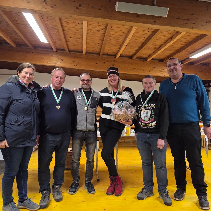 Eine Gruppe von Menschen steht zusammen, trägt Medaillen und lächelt. Eine Frau in der Mitte hält einen Korb. Sie sind in einem Raum mit Holzdecken und gelbem Boden.