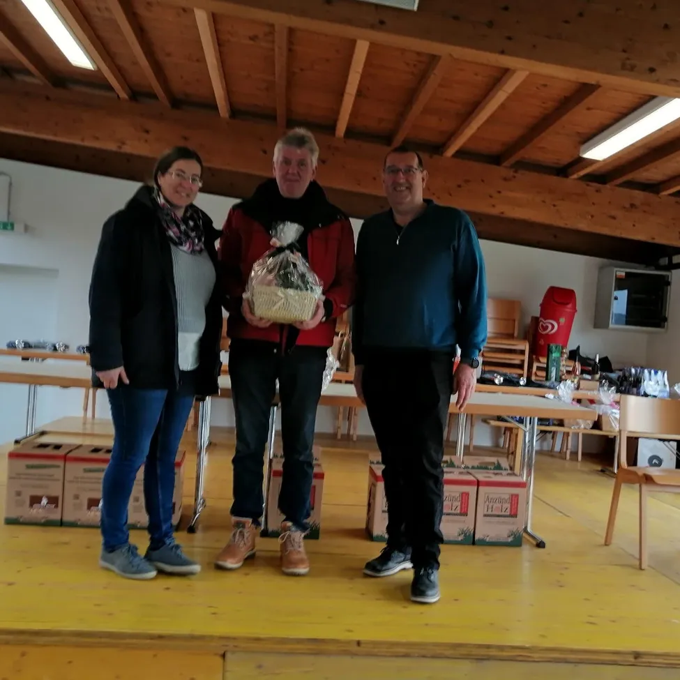 Drei Personen stehen auf einer Bühne, eine von ihnen hält einen Geschenkkorb. Hinter ihnen stehen Holztische und Stühle, und Kisten sind auf dem Boden platziert. Ein Feuerlöscher befindet sich auf der rechten Seite.