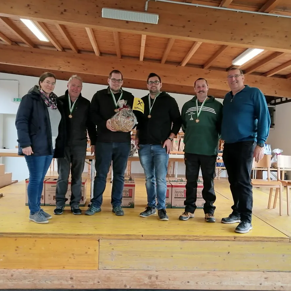 Fünf Personen stehen auf einer Bühne, tragen Medaillen und halten einen Brotbeutel. Sie feiern wahrscheinlich einen Sieg oder eine Leistung. Hinter ihnen befinden sich Holzstühle, Tische mit Kisten und eine Holzdecke.