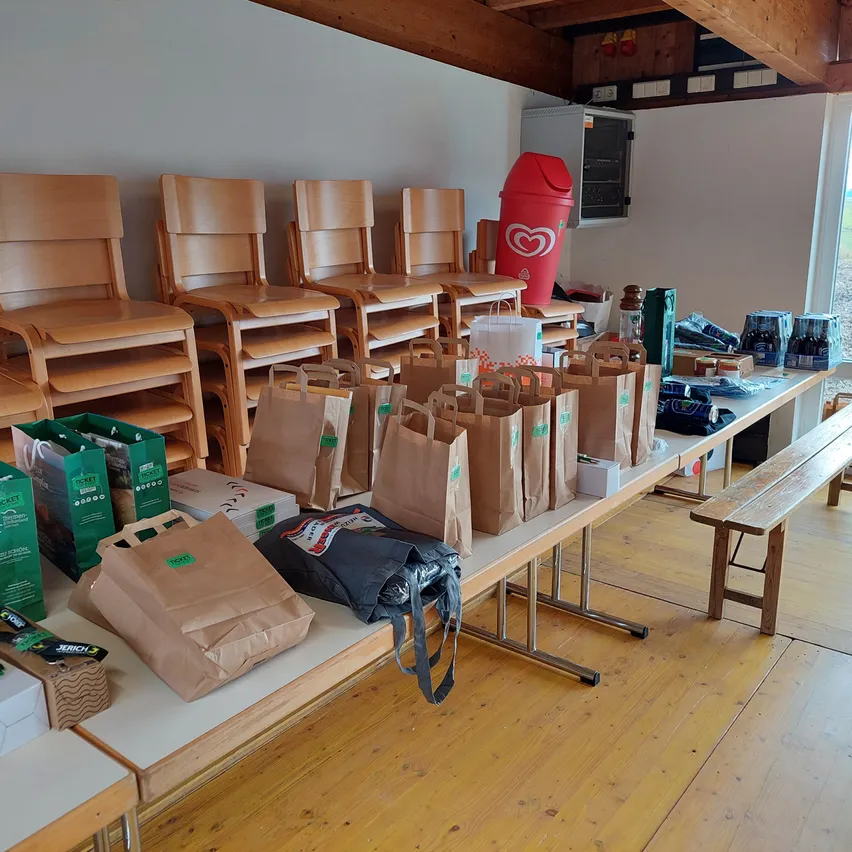 Ein Raum mit Holzboden und einer weißen Wand, der mehrere Holzstühle, einen Schreibtisch mit Taschen und Kisten, einen roten Mülleimer und einen Schrank mit Gegenständen darauf enthält.