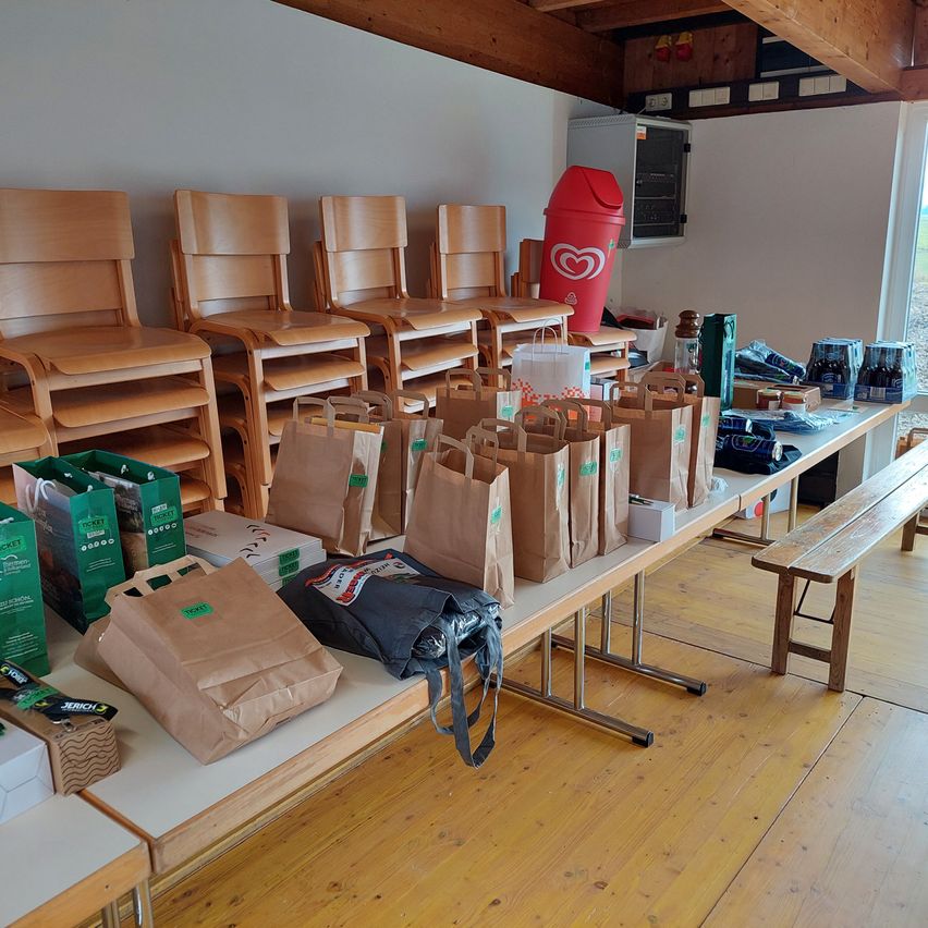 Ein Raum mit Holzboden und einer weißen Wand, der mehrere Holzstühle, einen Schreibtisch mit Taschen und Kisten, einen roten Mülleimer und einen Schrank mit Gegenständen darauf enthält.