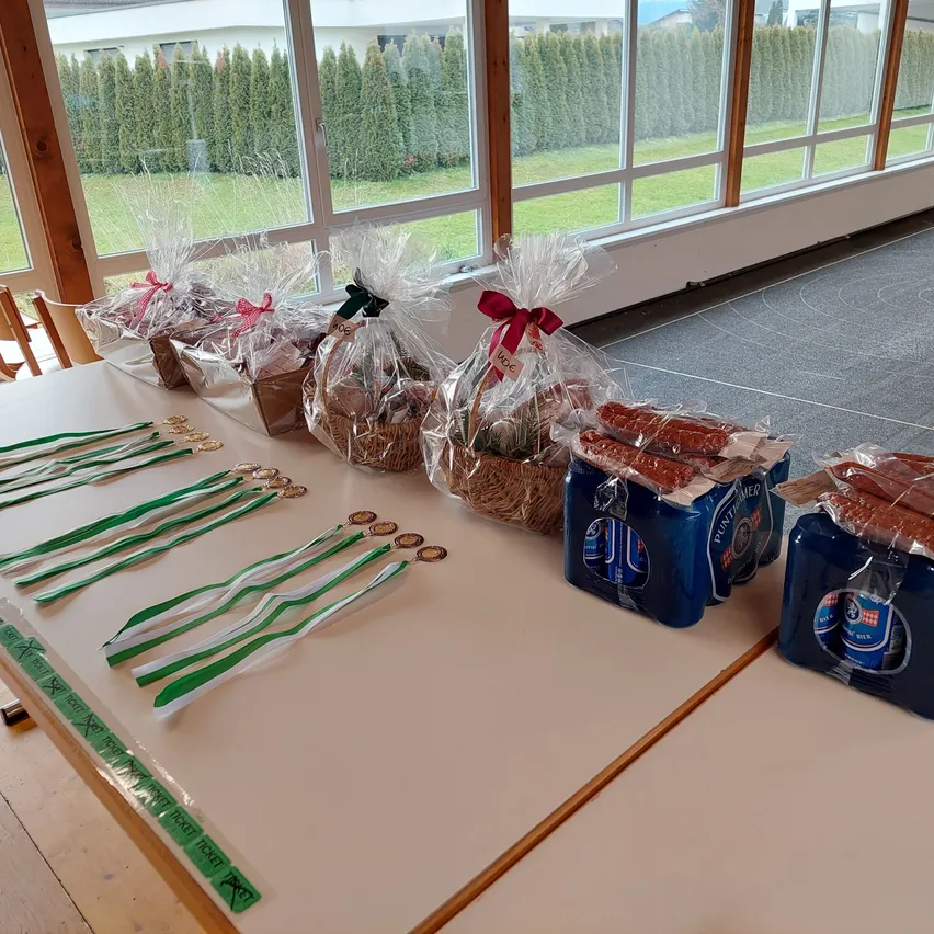 Ein Tisch zeigt verpackte Geschenke, Medaillen und Würste. Die Geschenke sind in Plastik mit roten Bändern verpackt. Medaillen sind in einer Reihe angeordnet und Würste in Plastikbehältern. Der Tisch steht vor einem großen Fenster.