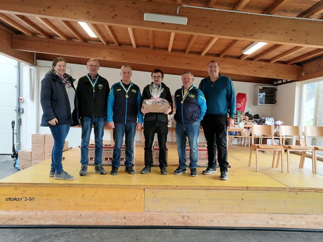 Sechs Personen stehen auf einer Bühne, möglicherweise bei einer Preisverleihung. Sie tragen alle Jacken und einige haben Medaillen. Ein Mann in der Mitte hält eine Schachtel mit einem Band. Hinter ihnen sind Stühle und Kisten zu sehen.