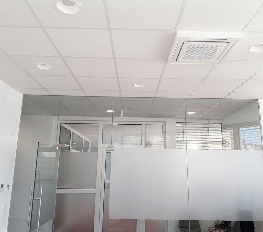 An office space features a white ceiling with recessed lighting and an air vent. The room has glass walls and doors, and blinds cover a window. A white partition separates the area.