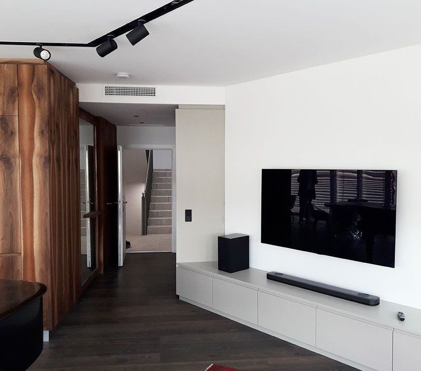 Modern living room with wooden flooring and white walls. A large flat-screen TV is mounted on the wall, with a speaker below. A wooden cabinet with a mirror is on the left.