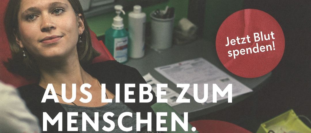 Eine Frau lächelt, während ihr der Blutdruck gemessen wird. Hinter ihr befindet sich eine medizinische Einrichtung mit einem Tisch, auf dem Dokumente und medizinische Utensilien liegen. Ein roter Kreis mit dem deutschen Text 'Jetzt Blut spenden' ist im Bild.